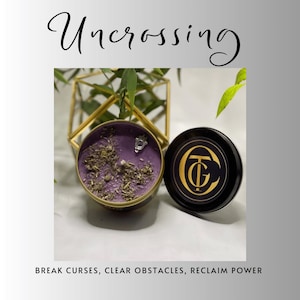 May include: A round, purple candle with dried herbs and a charm, next to a black lid with a gold logo. The candle is in a gold-rimmed tin. The word "Uncrossing" is written in cursive. Text at the bottom reads: BREAK CURSES, CLEAR OBSTACLES, RECLAIM POWER.