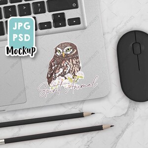 May include: A mockup of a laptop with a sticker of an owl with the text "Spirit Animal" and a sticker that says "JPG PSD Mockup". The laptop is on a white marble surface with two black pencils and a black computer mouse.
