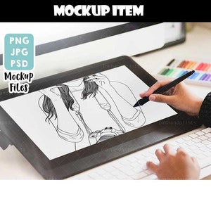May include: A black and white line drawing of a person holding a camera, displayed on a digital drawing tablet. The tablet is on a white desk with a keyboard and a set of colored pencils.