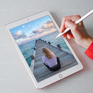 May include: A white tablet computer displaying a photo of a wooden pier extending into a body of water. A person is sitting on the pier, looking out at the water. The sky is blue with white clouds. The tablet is being used with a white stylus.
