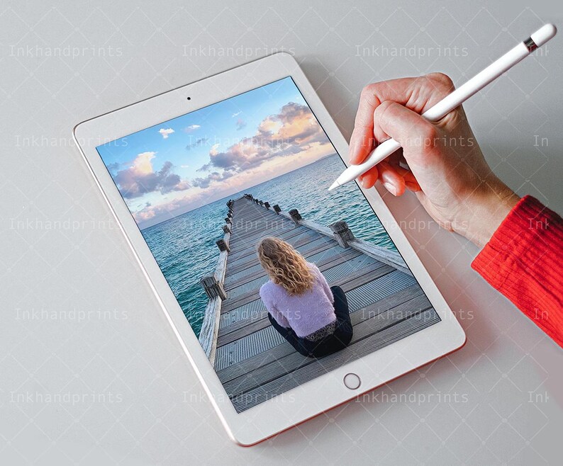 May include: A white tablet computer displaying a photo of a wooden pier extending into a body of water. A person is sitting on the pier, looking out at the water. The sky is blue with white clouds. The tablet is being used with a white stylus.