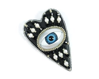 May include: A black heart-shaped brooch with a blue eye design. The brooch is embellished with white pearls, black beads, and silver trim. The eye has a detailed blue iris and a silver frame.