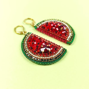 May include: A pair of watermelon slice earrings. The earrings have a red beaded design with a large, faceted red gem in the centre. The edges are trimmed with green, black, and gold beads, and have gold-coloured ear wires.