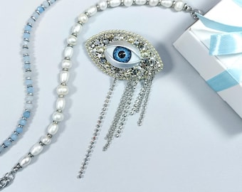 May include: An eye-shaped brooch with a blue iris, surrounded by silver and clear embellishments, and dangling silver chains. A white gift box with a light blue ribbon is in the background, along with two necklaces: one with blue beads and the other with white pearls.