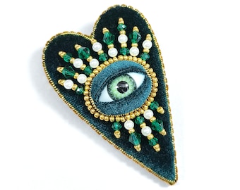 May include: A heart-shaped brooch in dark teal velvet, featuring an eye design at the center. The eye is surrounded by gold beads and embellished with green and white beads. The brooch has a gold beaded border.