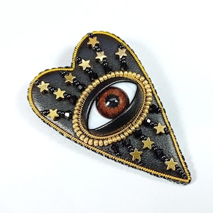 May include: A heart-shaped brooch with a black leather base, featuring a brown eye in the center. Gold stars and beads adorn the brooch, with a gold beaded border. The brooch has a unique, celestial design.