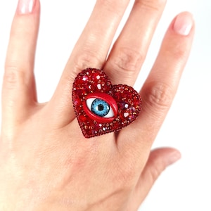 May include: A red heart-shaped ring with a blue eye design. The ring is covered in red crystals and features a detailed eye with blue iris and red lips. The ring is worn on a finger.