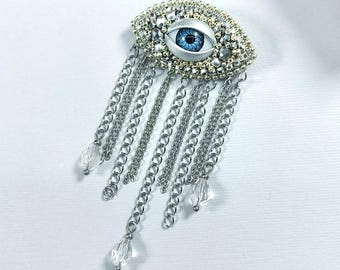 May include: A handcrafted brooch featuring a detailed eye design with a blue iris, surrounded by silver and gold beads. Silver chains with clear crystal drops cascade from the eye, creating a unique, elegant accessory.