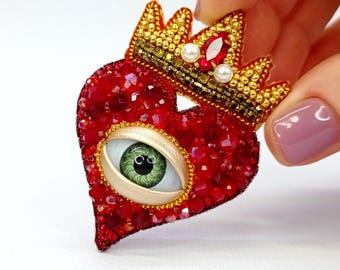 May include: A red heart-shaped brooch with a gold crown, adorned with red crystals, pearls, and a green eye. The brooch is intricately detailed, showcasing a unique and artistic design, perfect for adding a touch of personality to any outfit.