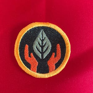 May include: Embroidered patch featuring a leaf held by two orange hands, set against a black background. The patch has a yellow border and is circular in shape. The background is red.