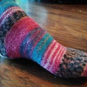 May include: A hand-knitted sock with a colorful striped pattern. The sock features bands of pink, teal, black, and white yarn. The heel and toe sections have a speckled design. The sock is displayed on a wooden surface.