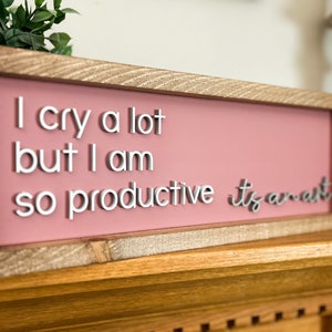 May include: A pink wooden sign with the text "I cry a lot but I am so productive it's an art" in white raised letters.