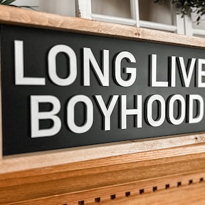 Könnte beinhalten: Ein gerahmtes Holzschild mit schwarzem Hintergrund und weißen erhabenen Buchstaben, auf dem "LONG LIVE BOYHOOD" steht.