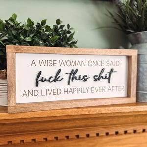 Könnte beinhalten: Holzgerahmtes Schild mit dem Text "A WISE WOMAN ONCE SAID fuck this shit AND LIVED HAPPILY EVER AFTER" in schwarzer Schrift. Das Schild steht auf einer Holzoberfläche mit Grün und einer Metallvase im Hintergrund.
