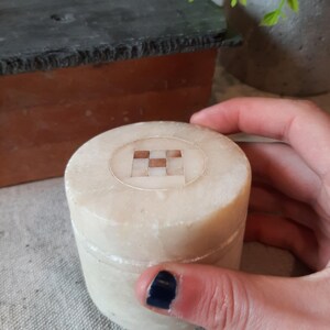 May include: A small, cylindrical, off-white stone box with a checkered design on the lid. The box is held by a hand with a dark blue painted nail. The box is likely used for storing small items.