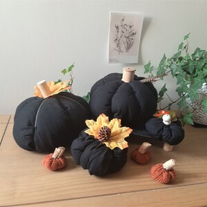 May include: A collection of fabric pumpkins in black and burnt orange. The larger black pumpkins feature wooden stems and decorative autumn leaves, while the smaller orange pumpkins have textured, ribbed surfaces and light-colored wooden stems.