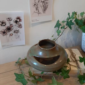 May include: A ceramic vase with a smiling face design, in shades of green and brown. The vase has a wide opening and sits on a wooden surface. Botanical art prints are visible on the wall behind the vase.