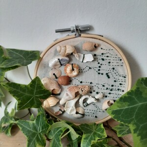 May include: An embroidery hoop art piece featuring various seashells and a stitched design in dark green thread. The shells are in shades of beige and brown, arranged on a light gray fabric. The wooden hoop is visible.