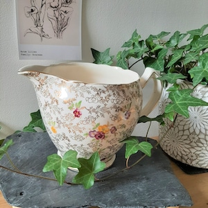 May include: Cream-colored vintage creamer with a floral and gold pattern. The creamer has a handle and a spout, and is decorated with small flowers in shades of purple, yellow, and red. The creamer is sitting on a dark gray surface.