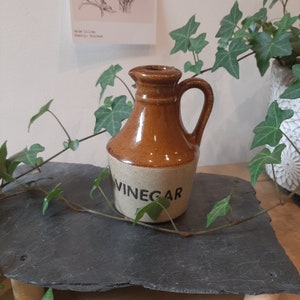 May include: A brown and beige ceramic vinegar jug with a handle. The word "VINEGAR" is printed in black on the beige base. The jug is displayed on a dark grey surface, with green ivy in the background.