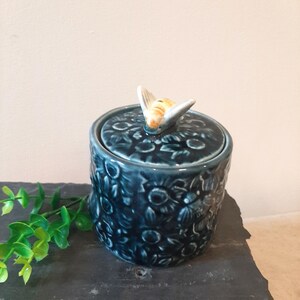May include: A blue ceramic honey pot with a bee-shaped lid. The pot is decorated with a floral pattern.