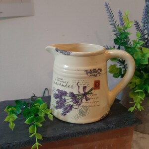 May include: Cream-coloured ceramic jug with a handle and spout. The jug is decorated with a lavender bouquet design, the word "Lavender", and postage stamp illustrations. The jug has a distressed, vintage appearance.