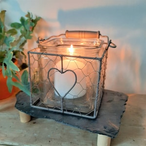 May include: A lit white candle in a clear glass jar, enclosed in a metal wire cage with a heart design. The jar sits on a dark grey slate-like surface with wooden legs. A wooden handle is on top. The candle is lit.