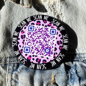 Könnte beinhalten: Ein runder schwarz-weißer Aufkleber mit einem rosa-schwarzen Leopardenmuster im Hintergrund. Der Aufkleber hat einen QR-Code in der Mitte und den Text "SCAN ME" wiederholt am Rand.