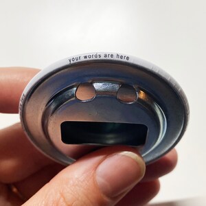 May include: A white metal bottle opener with a silver metal ring and a cutout in the shape of a bottle cap. The words "your words are here" are printed around the edge of the opener.