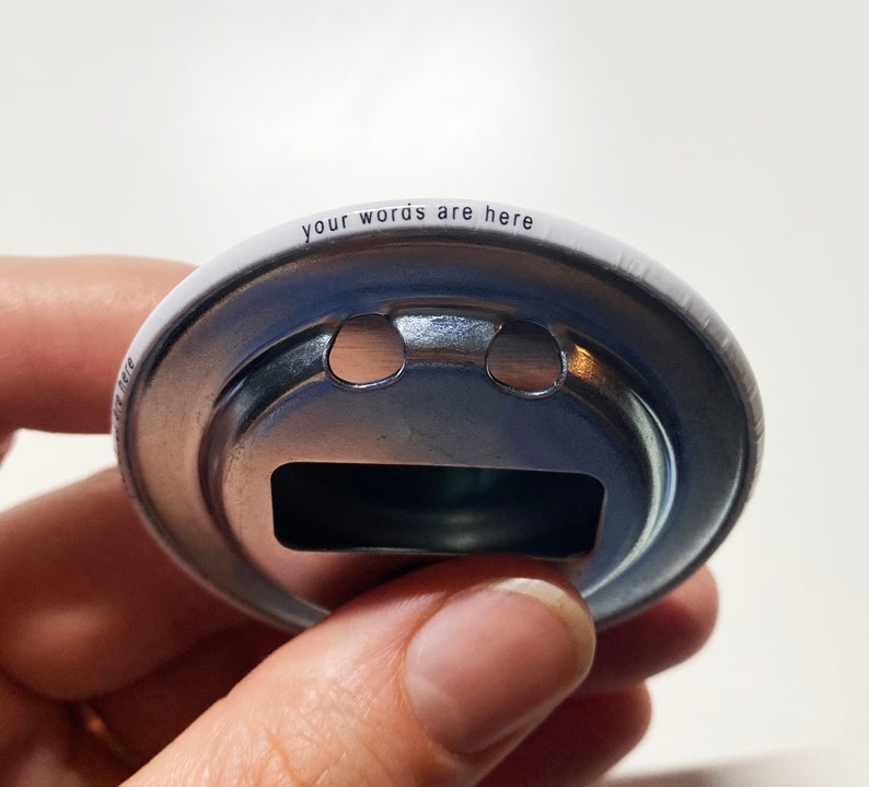 May include: A white metal bottle opener with a silver metal ring and a cutout in the shape of a bottle cap. The words "your words are here" are printed around the edge of the opener.