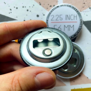 May include: The back of a 2.25 inch (56 mm) button with a bottle opener attached. The button is silver with a black rim. The button is being held by a hand.