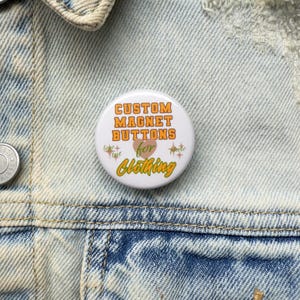 May include: A white custom magnet button with orange text that reads "CUSTOM MAGNET BUTTONS for Clothing". The button has a heart and starburst graphic. The button is on a denim jacket.
