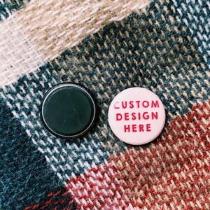 May include: Two round buttons, one black and one pink. The black button is blank. The pink button has the text "CUSTOM DESIGN HERE" in pink letters.