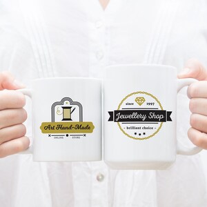 May include: Two white ceramic mugs with black and gold logos. The mug on the left has a logo that says "Art Hand-Made" with a sewing needle and thread design. The mug on the right has a logo that says "Jewellery Shop" with a diamond design and the text "since 1997".