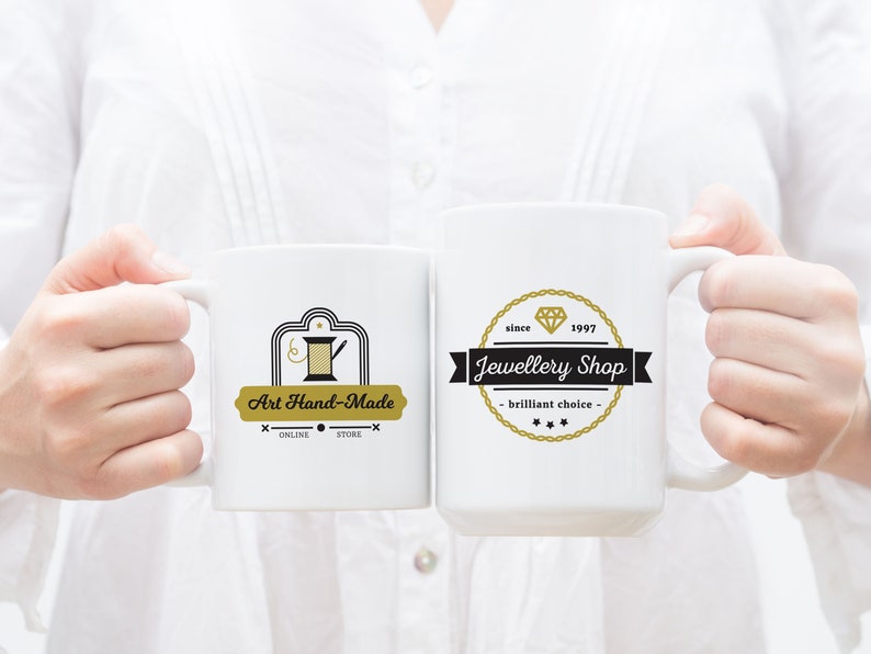 May include: Two white ceramic mugs with black and gold logos. The mug on the left has a logo that says "Art Hand-Made" with a sewing needle and thread design. The mug on the right has a logo that says "Jewellery Shop" with a diamond design and the text "since 1997".
