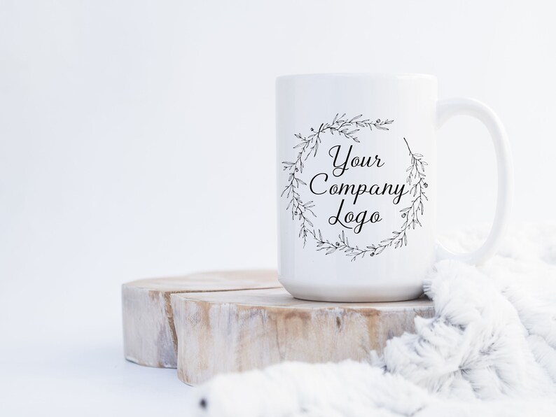 May include: White ceramic mug with a black floral wreath design and the text "Your Company Logo". The mug is sitting on a wooden slice and a white fuzzy blanket.