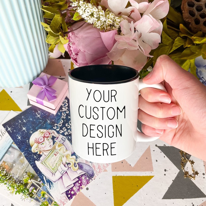 May include: White ceramic mug with a black rim and handle. The mug has the text "YOUR CUSTOM DESIGN HERE" printed in black on the front.