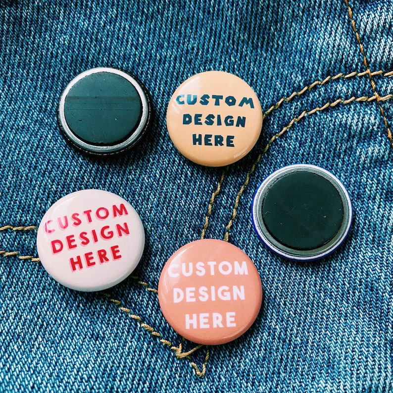 May include: Four round buttons with a denim background. Two buttons are peach colored with the text "CUSTOM DESIGN HERE" in black and red. One button is white with the text "CUSTOM DESIGN HERE" in black and red. One button is black with a silver rim.