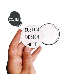 May include: A white 2.25 inch button with the text "CUSTOM DESIGN HERE" printed in black.
