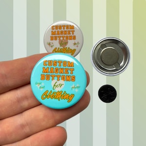 Op de afbeelding: Lichtblauwe en witte aangepaste magneetknoppen met de tekst "CUSTOM MAGNET BUTTONS for Clothing". Een magneet en een zwarte magneet zijn ook afgebeeld.