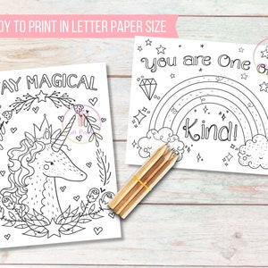May include: Two black and white coloring pages with the phrases "Stay Magical" and "You are One of a Kind!" are displayed. The first features a unicorn, and the second a rainbow. A bundle of pencils rests on the wooden surface.