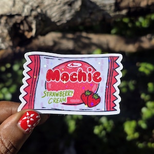 Puede incluir: Una pegatina con un diseño de envoltorio de caramelo mochi de fresa. El envoltorio es rojo con la palabra "Mochie" en rojo y "Strawberry Cream" en verde. Dos fresas están en la parte inferior derecha. La pegatina tiene un efecto brillante.