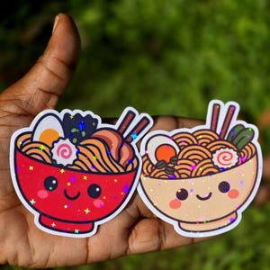 May include: Two ramen bowl stickers. One is red, the other is beige. Both have smiling faces, noodles, eggs, and chopsticks. The stickers have a holographic, glittery effect.
