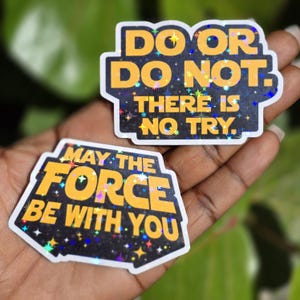 Może przedstawiać: Dwie wycinane naklejki z czarnym tłem i holograficznym wzorem gwiazd. Górna naklejka zawiera żółty napis DO OR DO NOT. THERE IS NO TRY. Dolna naklejka zawiera żółty napis MAY THE FORCE BE WITH YOU.