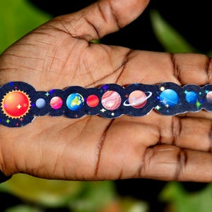 May include: A colorful sticker featuring the solar system, including the sun and planets. The sun is depicted with red and orange hues, while the planets showcase various colors and details. The sticker is held in a hand.