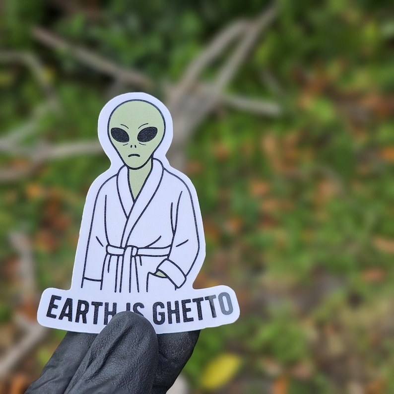 May include: A sticker depicting a green alien in a white robe, with large black eyes and a sorrowful expression. The text "EARTH IS GHETTO" is written in white on a curved banner beneath the alien.