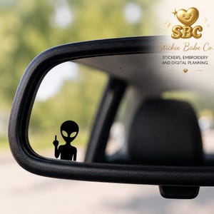 Può includere: Un adesivo in vinile nero raffigurante una figura aliena che fa il gesto del dito medio, applicato sulla superficie di uno specchietto laterale dell'auto. L'alieno ha una grande testa ovale e occhi scuri, posizionato nell'angolo in basso a sinistra.