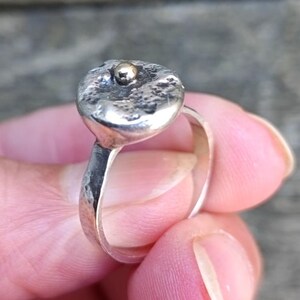 Op de afbeelding: Een zilveren ring met een getextureerde, onregelmatige bovenkant en een kleine goudkleurige bol in het midden. De ringband is dun en glad. De ring wordt door een persoon vastgehouden.