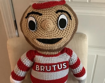 May include: A crocheted plush toy of Brutus Buckeye, the mascot of Ohio State University. The toy is wearing a red and white striped shirt with the word "BRUTUS" on the front, a white hat with a red "O" on it, and a white towel with a red "O" on it.