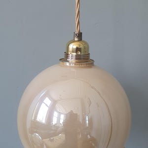 Op de afbeelding: Een hanglamp in vintage-stijl met een bolvormige, crèmekleurige glazen kap. De kap heeft een geschulpte onderrand en wordt opgehangen door een goudkleurige metalen armatuur en een gedraaid stoffen snoer. De lamp is een decoratief element voor in huis.